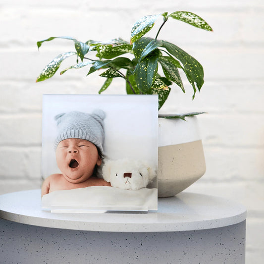 Acrylic desk print of a newborn baby wearing a knit hat and yawning, displayed on a round white side table next to a potted plant.