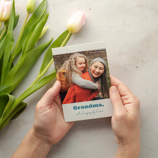 Flat photo card held in hands featuring a child hugging an older adult, with “Grandma, A Legacy of Love” text beside pink tulips.