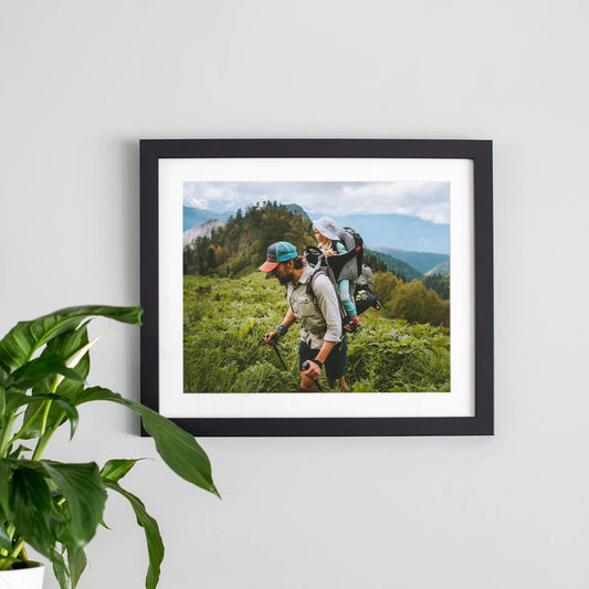 Framed print with white mat and black frame showing person hiking with child in backpack across green mountain landscape, horizontal orientation, displayed on wall.