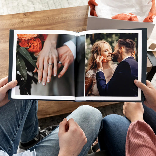 Hardcover wedding photo book held open, showing a two-page spread with a close-up of hands and a couple sharing a moment