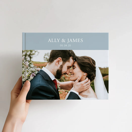 8.5x11 hardcover photo book with a personalized wedding cover, showing a couple embracing outdoors, held in one hand against a neutral background.