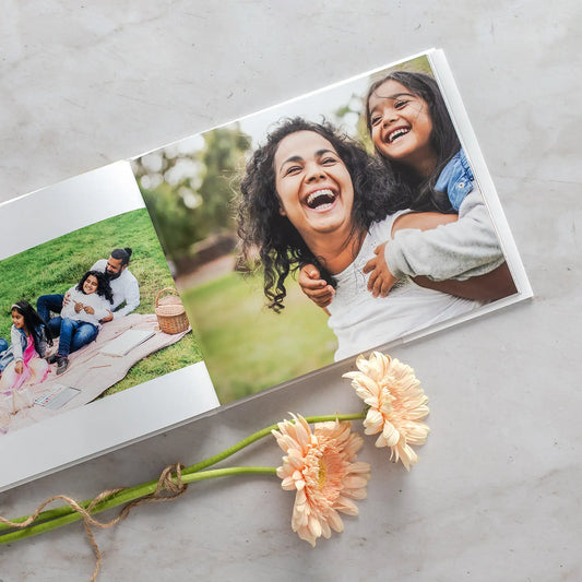 Hardcover photo book open to full-bleed spread with family picnic scene and a close-up of a person giving a child a piggyback, styled with flowers.