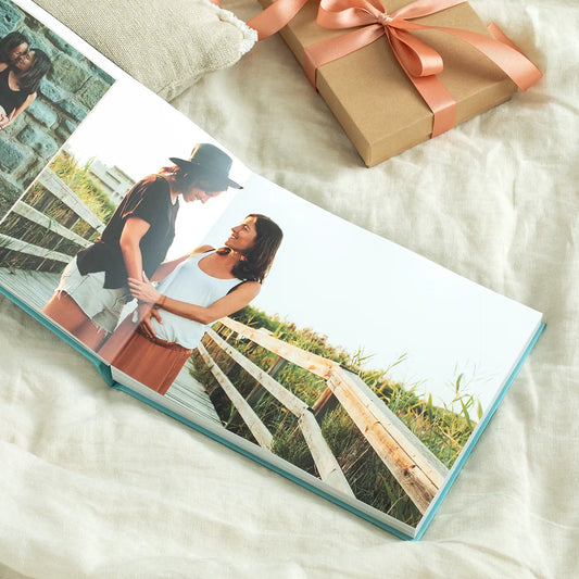 Layflat photo book open to a couple standing together on a wooden boardwalk, styled with a wrapped gift box nearby