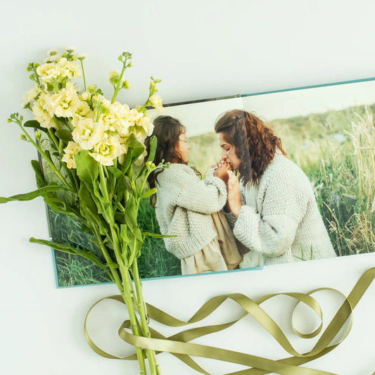 Layflat photo book showing a parent and child outdoors in a grassy field, styled with fresh flowers and ribbon on a white surface.