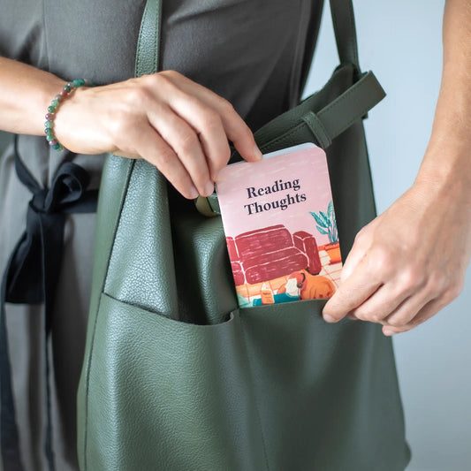 A person placing a small memo book titled “Reading Thoughts” into a green tote bag, shown at chest level.