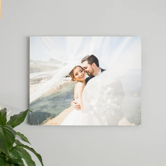 Metal wall print featuring a bride and groom embracing near a coastal cliff with flowing veil, mounted on a light gray interior wall.