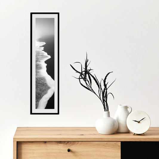 Vertical panoramic print of a black and white aerial shoreline with waves meeting sand, displayed in a slim black frame above a wooden cabinet.