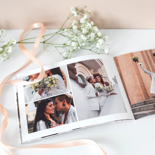 Open softcover photo book displaying a wedding collage spread with multiple couple portraits, styled with ribbon and small white flowers on a tabletop