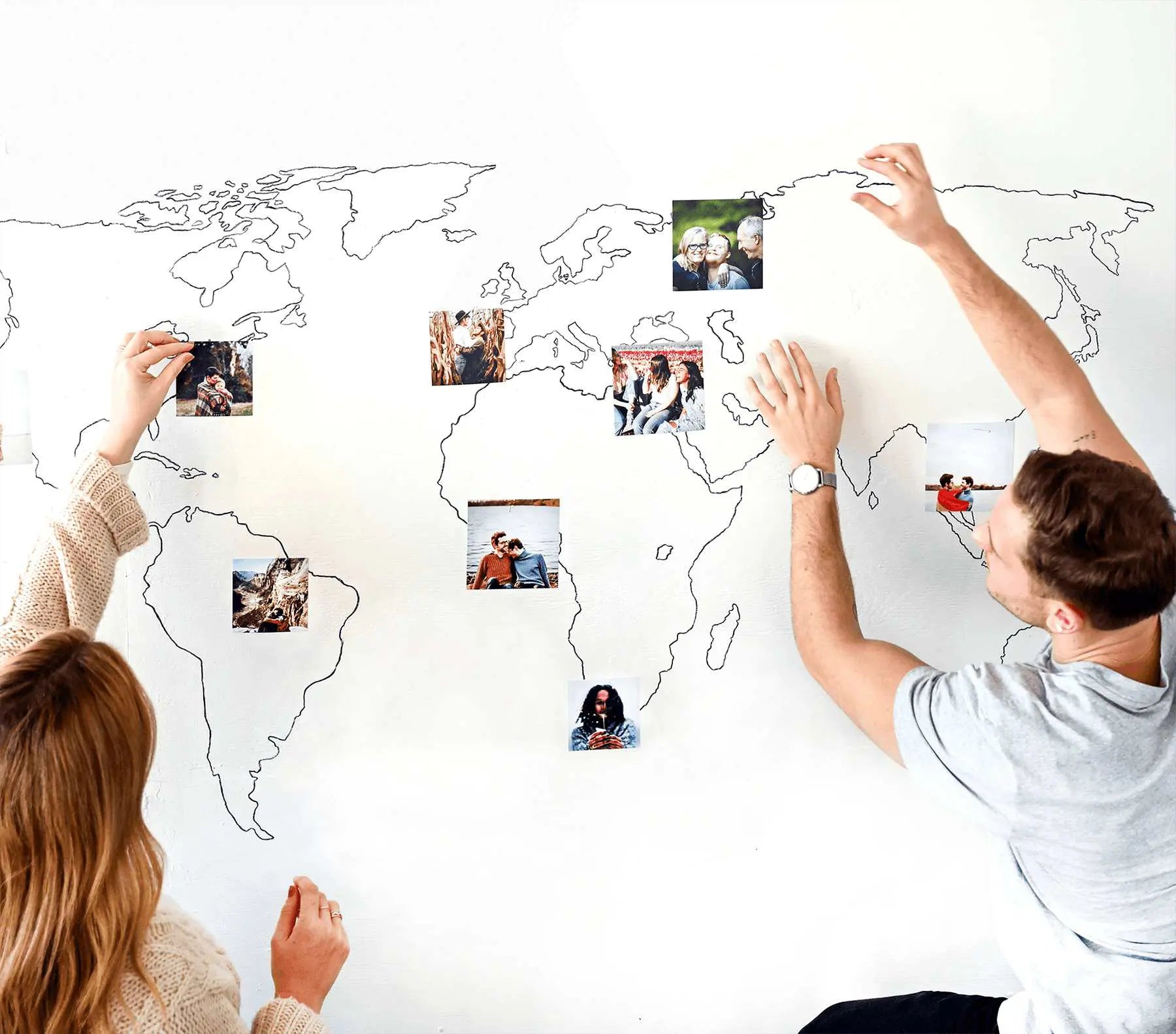 Two people placing small square photo prints on a wall with a minimalist world map outline, arranging travel photos across different continents.