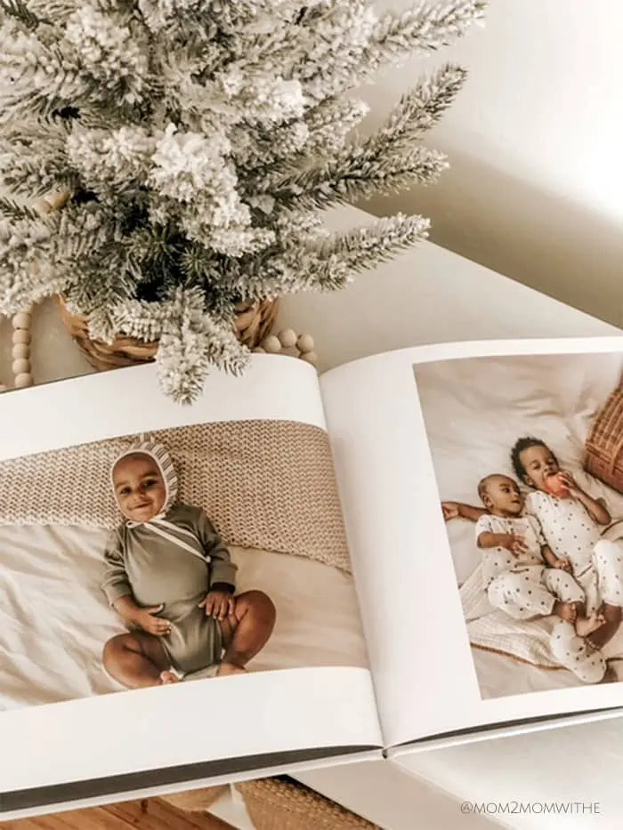 Open photo book displaying baby photos on thick white pages beside a small decorative tree and wooden beads on a light tabletop.