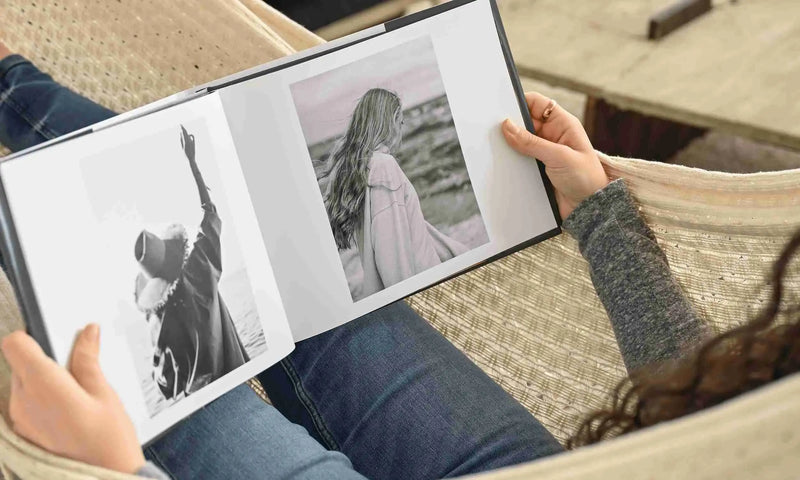 Person sitting outdoors holding an open hardcover photo book with black-and-white portraits printed on thick white pages while relaxing on a woven lounge chair.