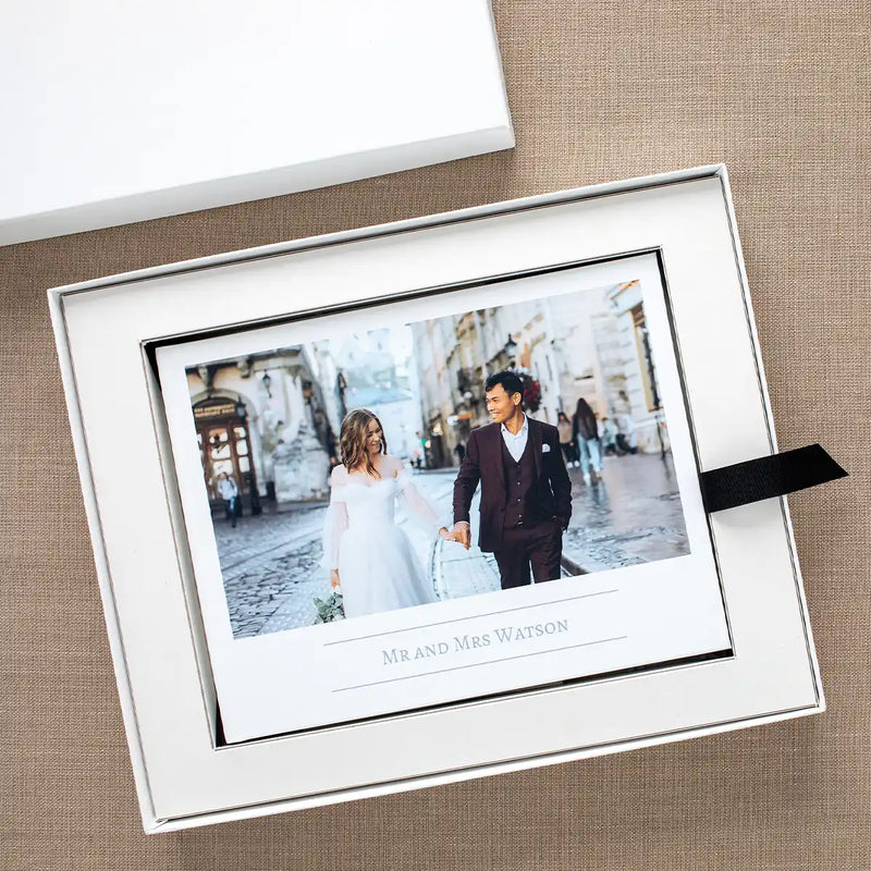 Layflat wedding photo book displayed inside white keepsake box with black ribbon, featuring a couple on the cover photo and custom title text.
