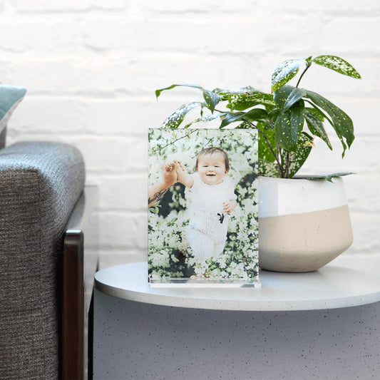 Acrylic desk print of a smiling baby standing among white blossoms, displayed on a round side table beside a modern plant pot.
