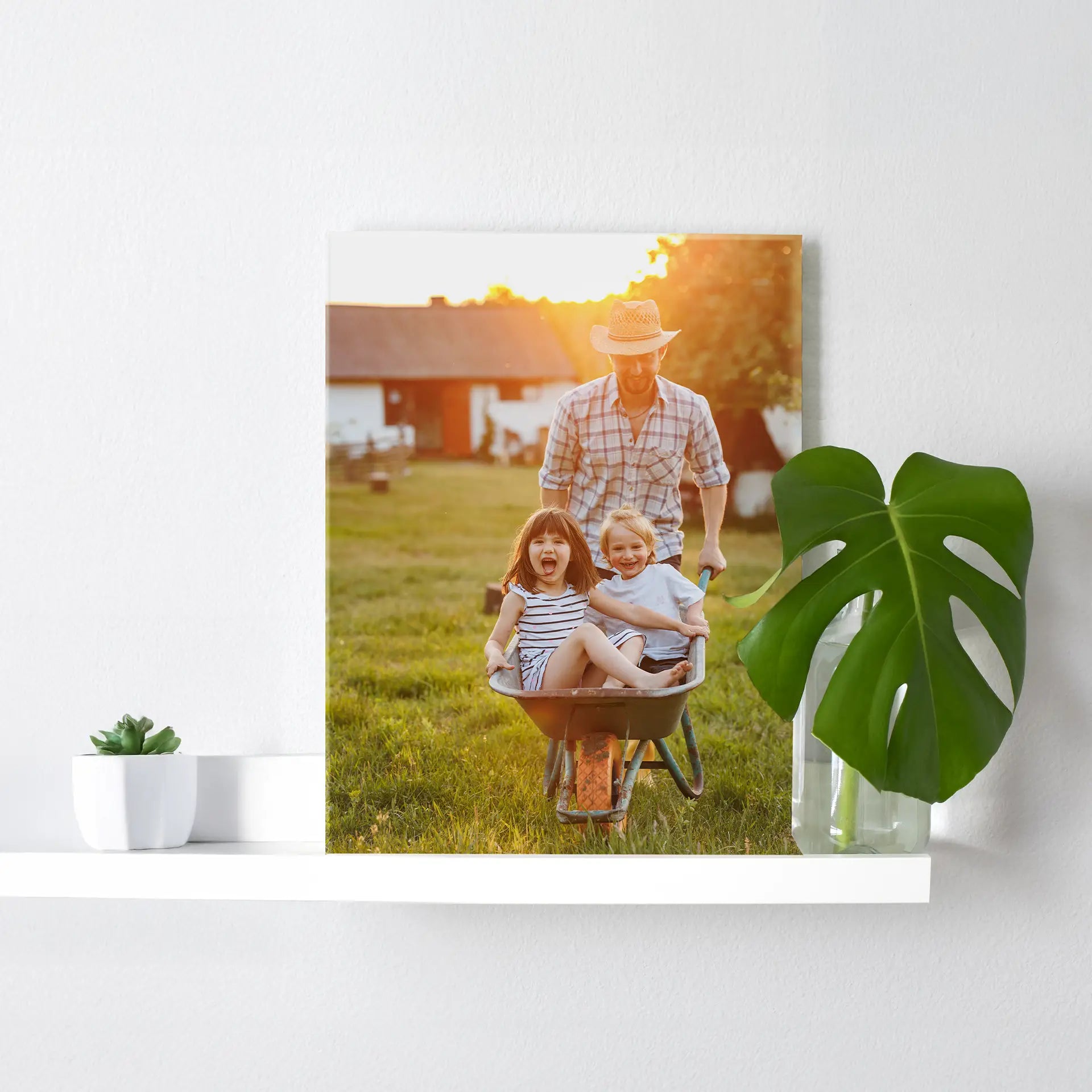 Acrylic wall decor print featuring an adult pushing two children in a wheelbarrow outdoors at sunset, displayed on a white shelf with a potted plant.