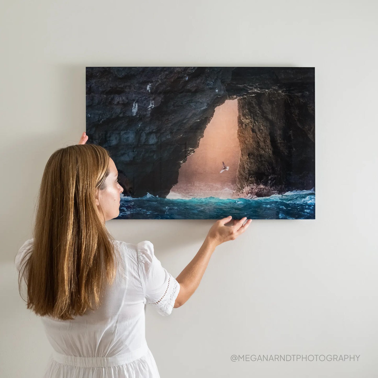 Acrylic wall decor print showing ocean waves inside a rocky cave opening with sunlight streaming through, being hung on a white wall.