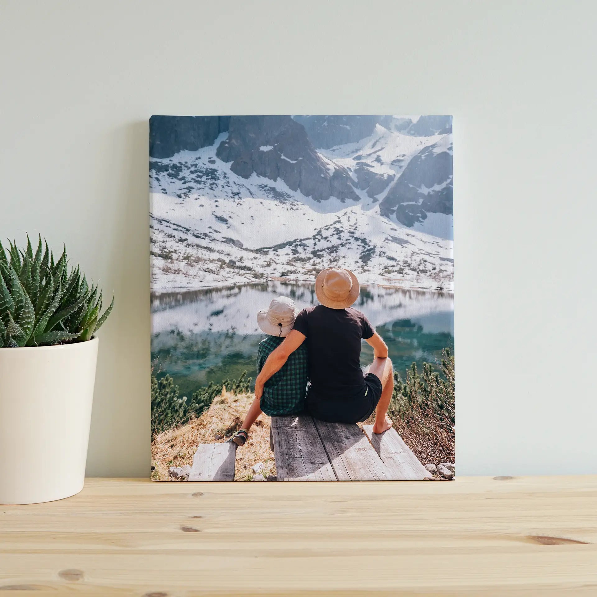 Canvas desk print of an adult and child sitting on a wooden dock overlooking a mountain lake, displayed beside a potted succulent.