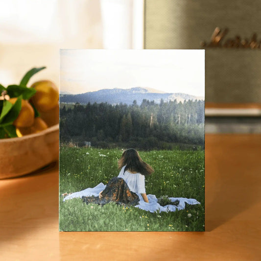 Canvas desk print featuring a woman sitting on a blanket in a green meadow with mountains in the distance, styled on a wooden table.