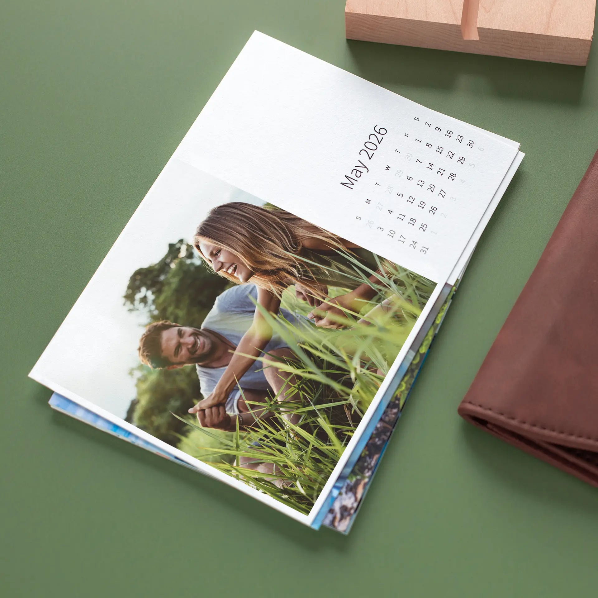 Stacked desk calendar pages showing a smiling couple lying in tall green grass, with the month and date grid visible at the top.