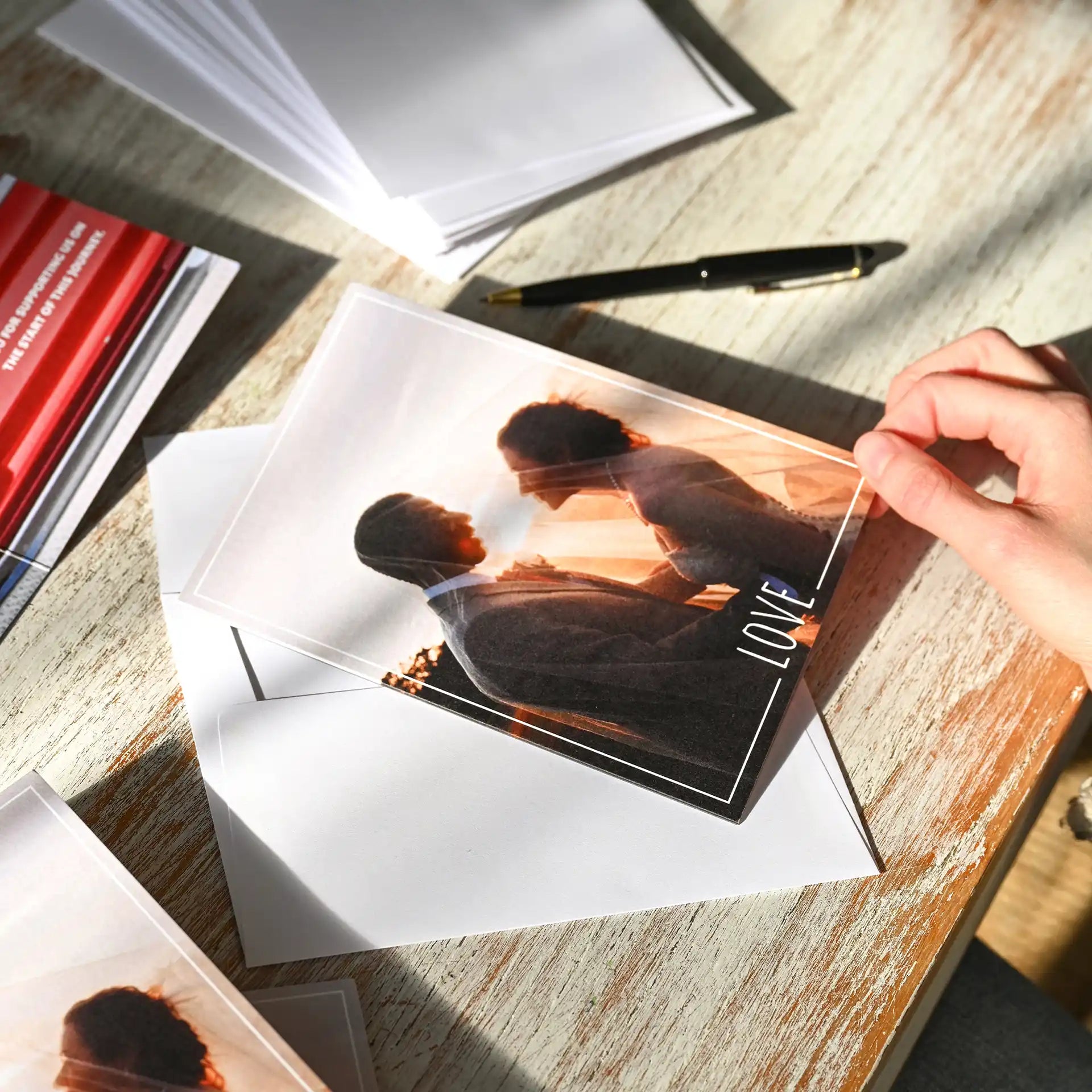 Folded photo card with translucent overlay showing silhouetted couple at sunset and white “LOVE” text, held above envelope on wooden table.