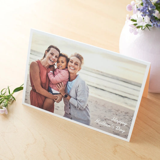 Folded photo card featuring three people smiling on a beach with “Happy Mother’s Day” text, displayed on a wooden surface with flowers nearby.