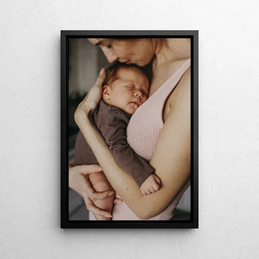 Framed canvas print with black frame featuring close-up of a person holding a sleeping newborn, vertical orientation, soft indoor lighting, displayed on neutral wall.