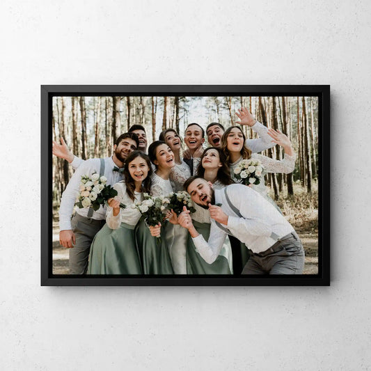 Framed canvas print with black frame featuring group portrait of people in wooded setting holding bouquets, wide landscape orientation, displayed on wall.