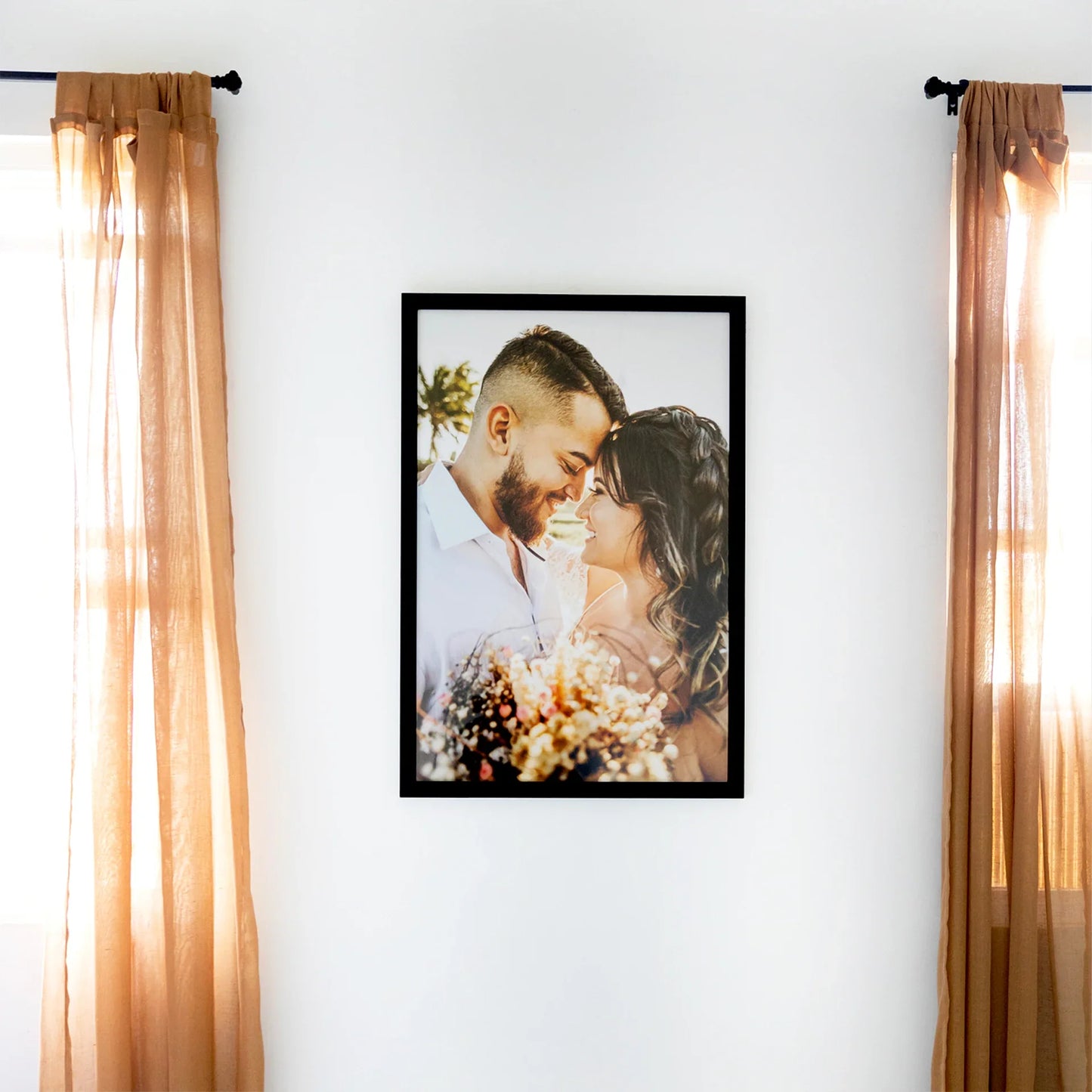 Black framed vertical photo print of a smiling couple embracing at sunset, displayed on a white wall between sheer curtains.