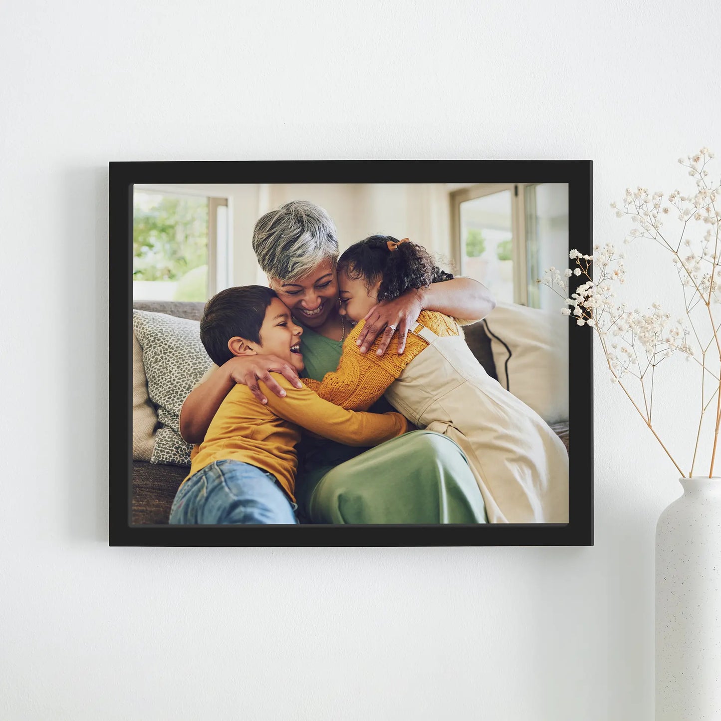 Black framed horizontal photo print of an older woman hugging two children on a couch in a bright living room, mounted on a white wall beside a vase with dried flowers.