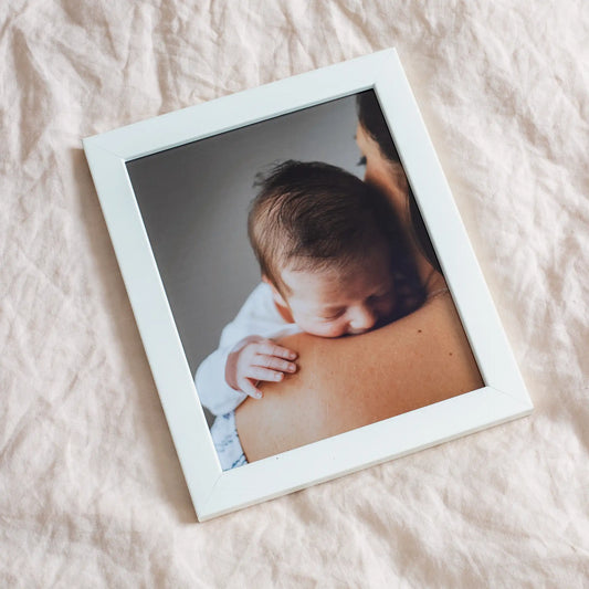 Framed photo print with white border frame showing a close-up portrait of a mother holding a sleeping newborn baby indoors.