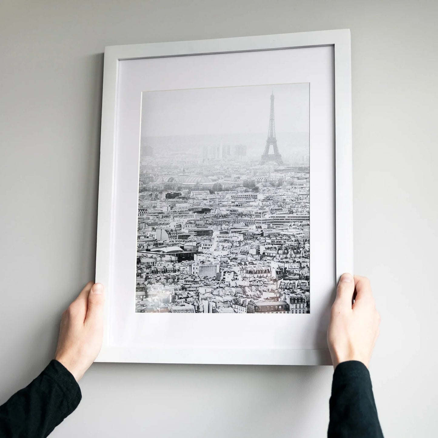White matted square framed black and white aerial photo of Paris rooftops with the Eiffel Tower visible in the distance, held against a gray wall.