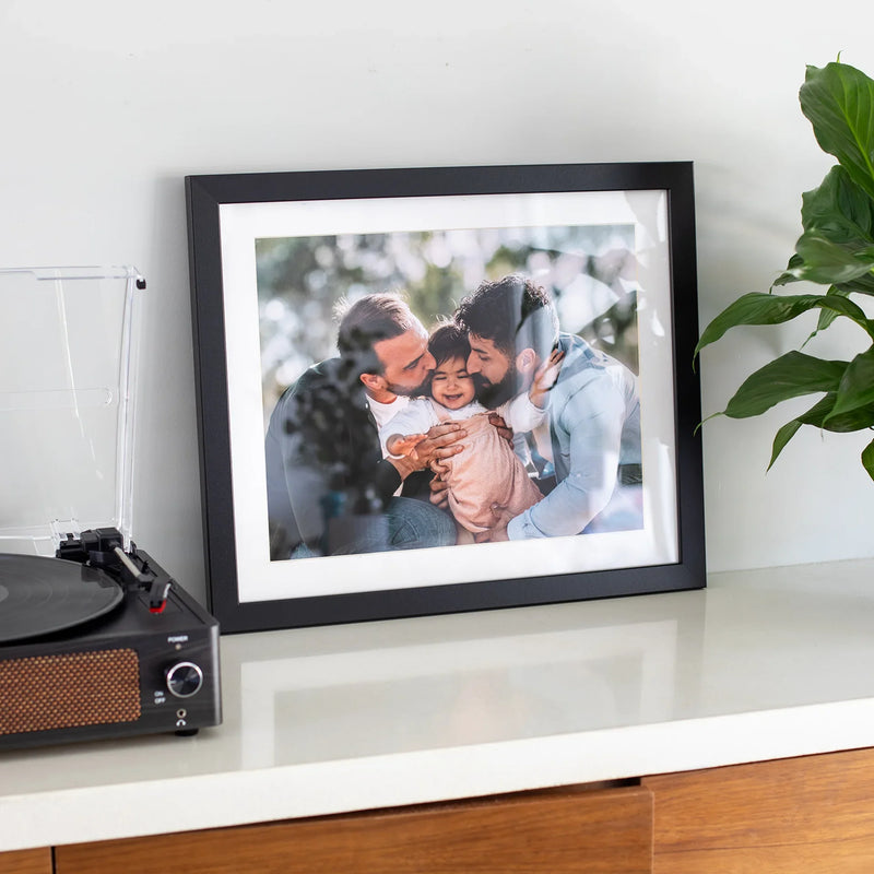 Black matted horizontal framed photo print of two adults hugging a child outdoors, displayed on a white shelf beside a record player and plant.
