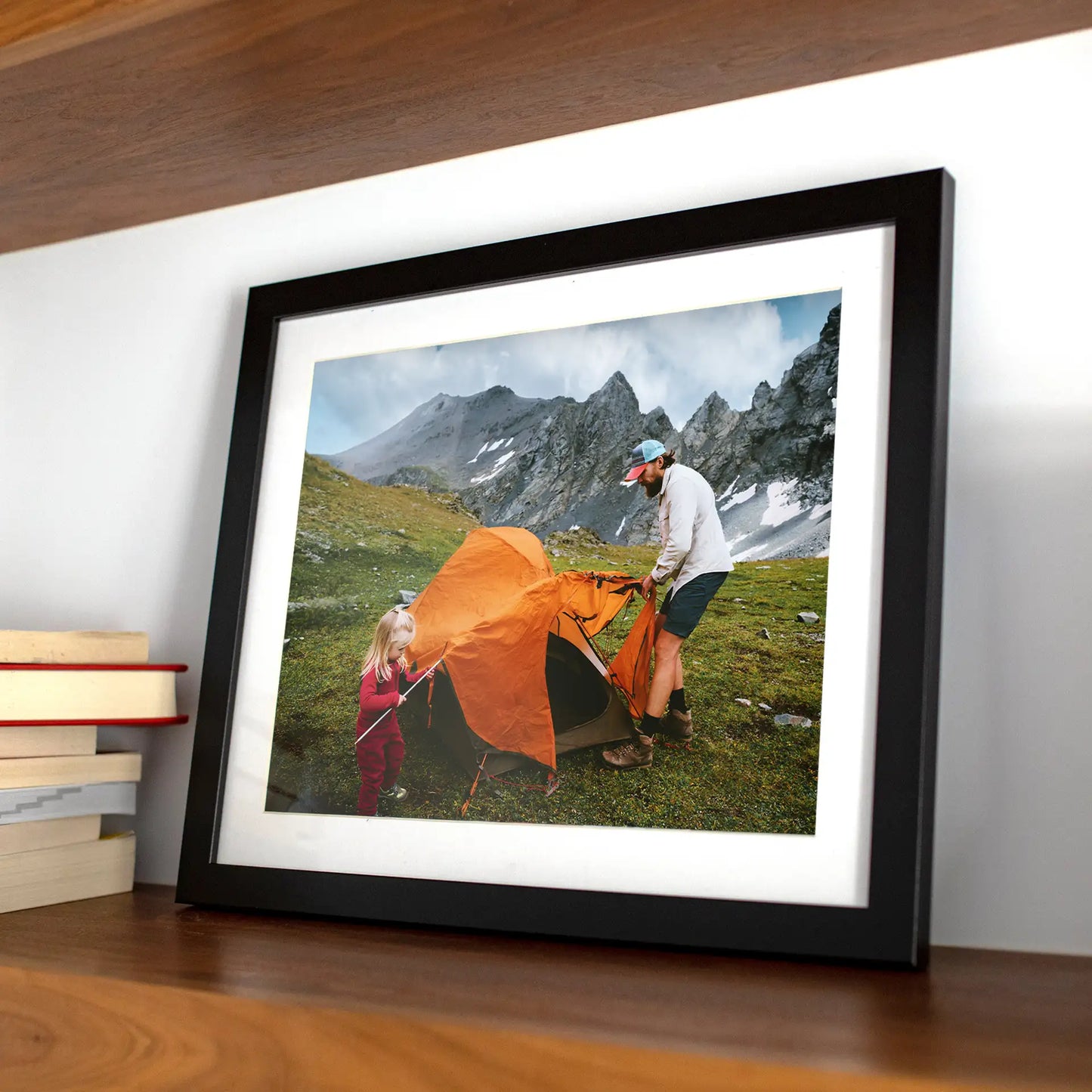 Black matted horizontal framed photo of an adult and child setting up an orange tent in a grassy mountain landscape with rocky peaks behind them.