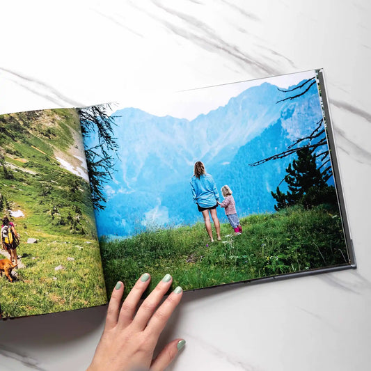 Hardcover photo book open to a full-bleed spread showing a parent and child standing in a grassy mountain landscape, viewed from behind
