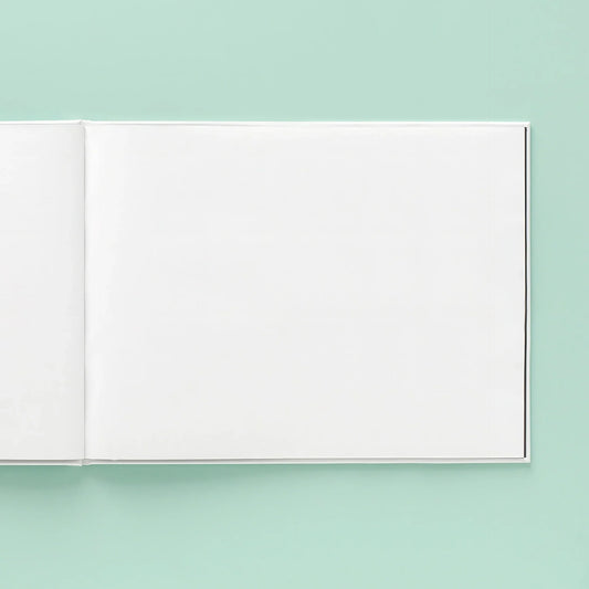 Blank layflat photo book with smooth matte pages and hinge binding, open at wide angle on teal surface highlighting crease-free panoramic layout.