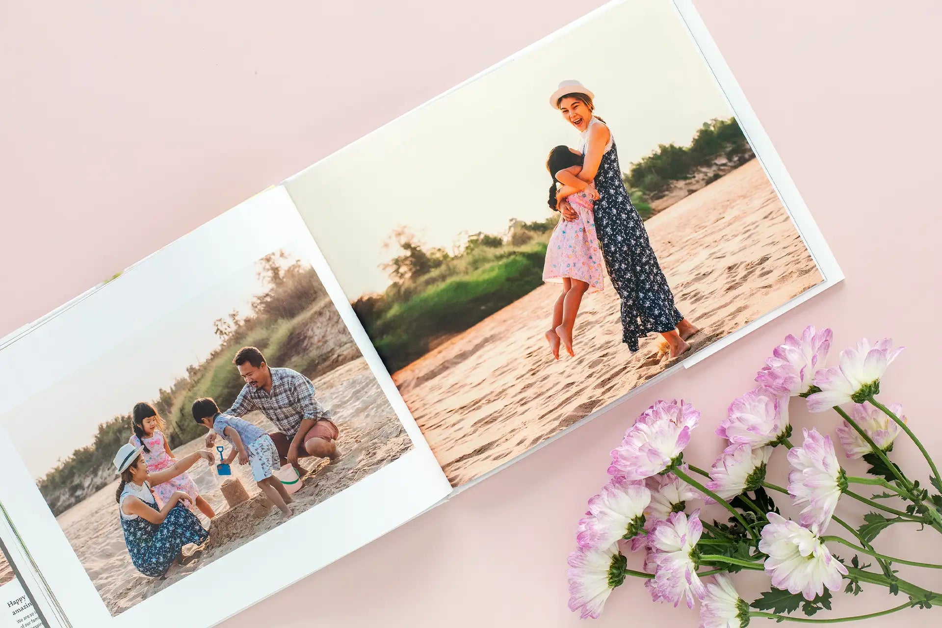 Hardcover photo book open to beach family photos with parents and children playing in sand, styled with pink flowers on soft pink surface.