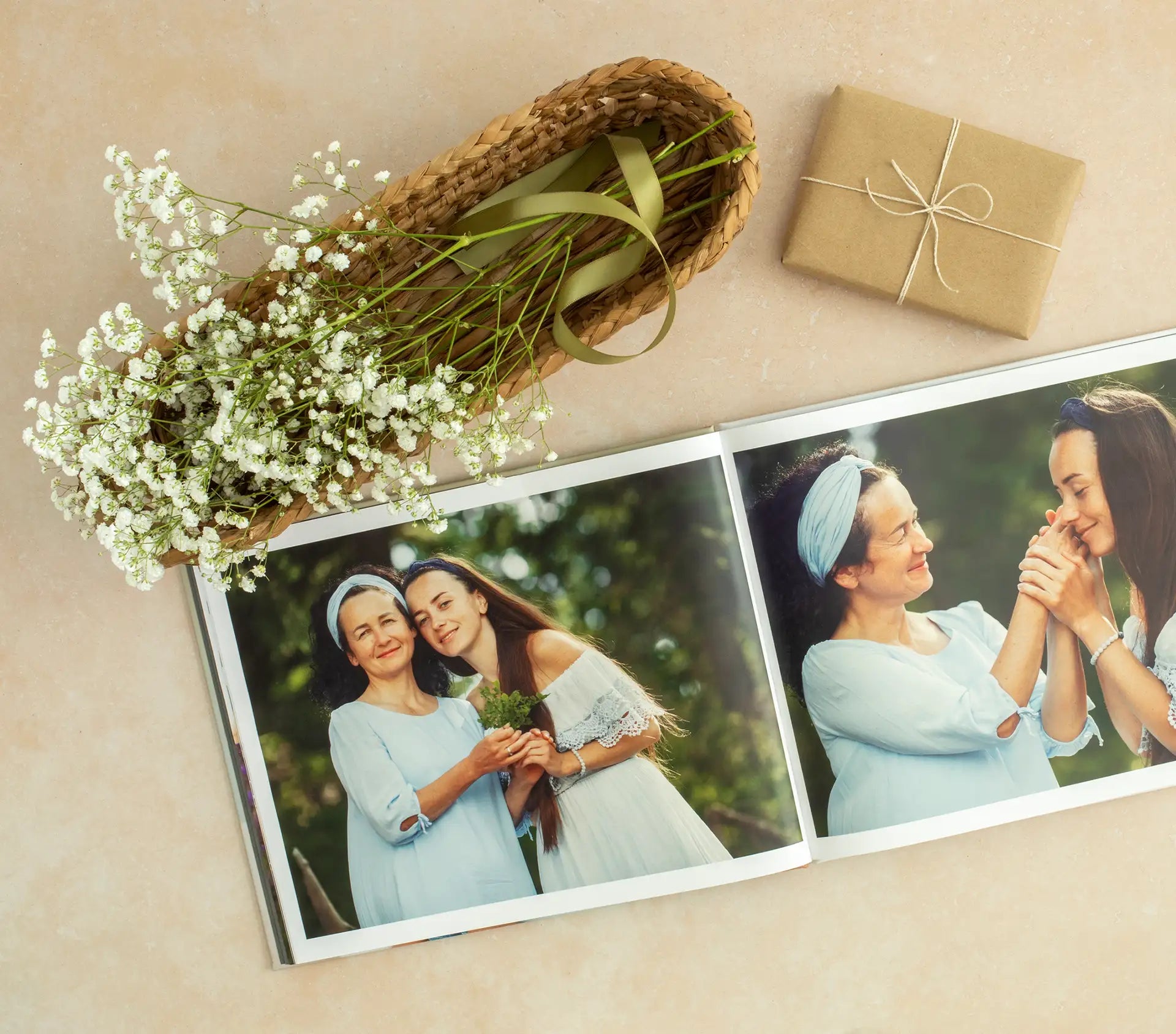 Hardcover photo book open to portrait photos of mother and daughter outdoors, styled with wrapped gift and small white flowers on neutral background.