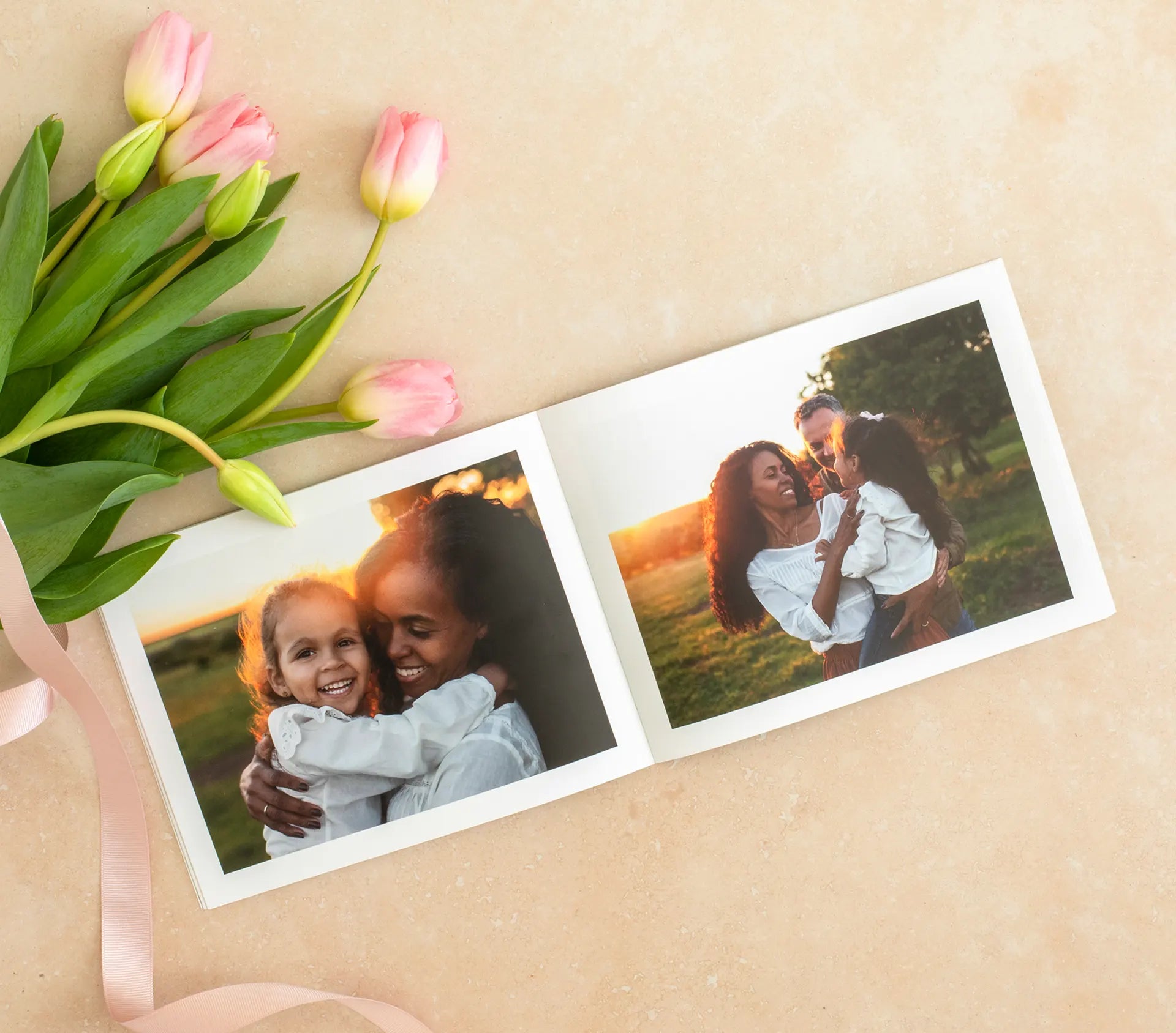 Softcover photo book open to two-page spread showing mother and child hugging outdoors at sunset, styled with pink tulips on neutral background.