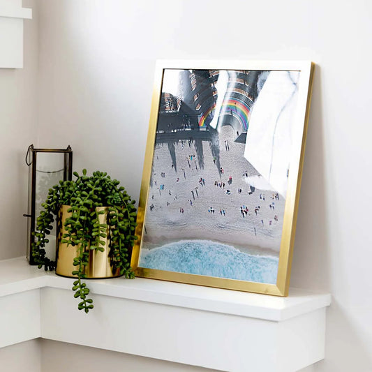 Gold framed large-format vertical photo print of an aerial beach scene with people, ocean shoreline, and a rainbow-colored architectural installation, displayed on a white corner shelf.