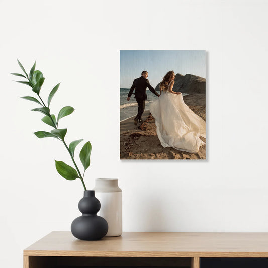 Maple wood print with natural grain showing couple walking along beach shoreline, vertical orientation, displayed above wooden console with decorative vases and plant.