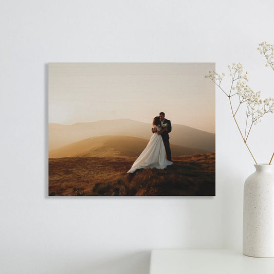 Maple wood print with warm wood grain featuring couple embracing on hillside at sunset, horizontal orientation, displayed on wall beside minimalist vase.