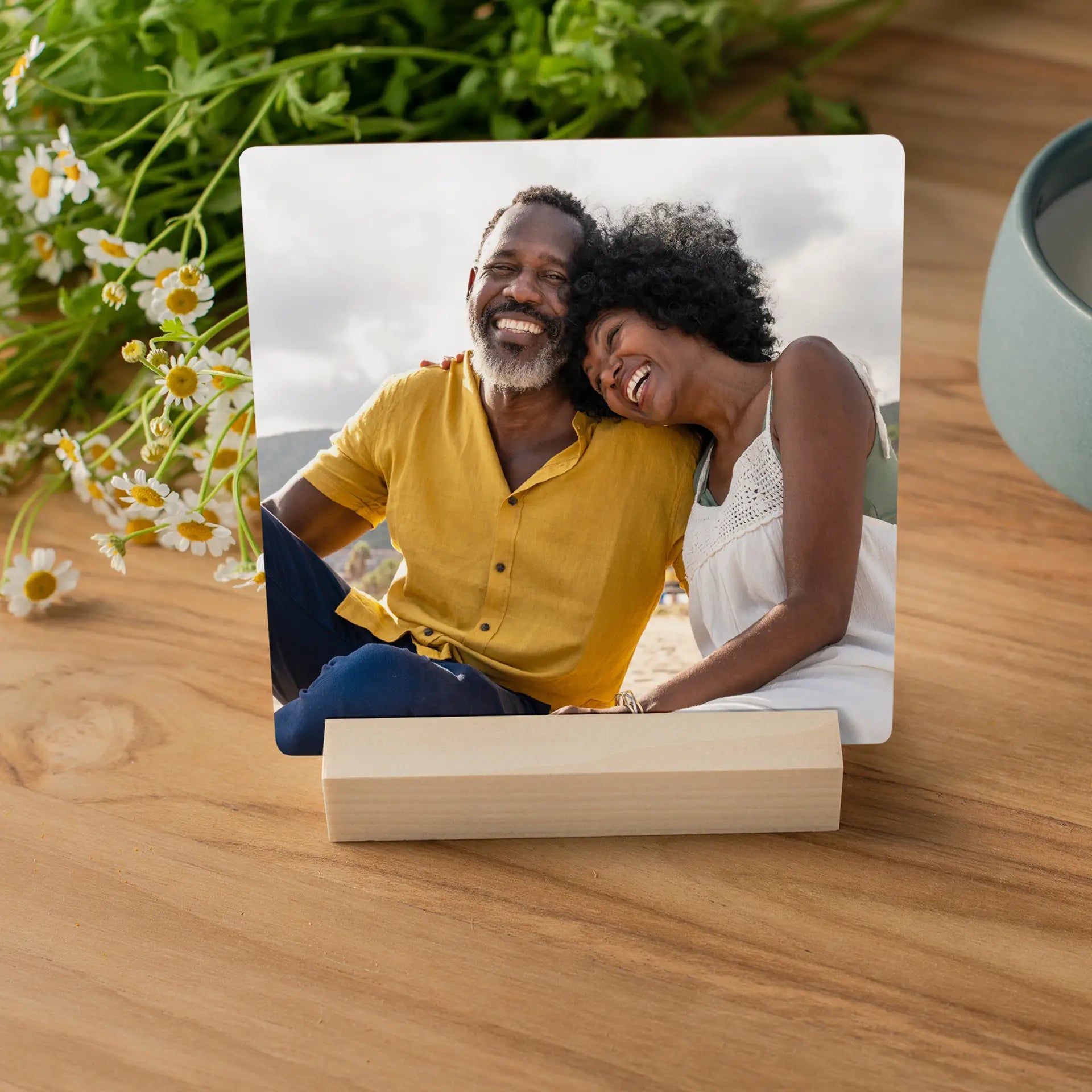 Metal desk print featuring a smiling couple leaning together outdoors, displayed in a light wood block on a wooden table with flowers nearby.