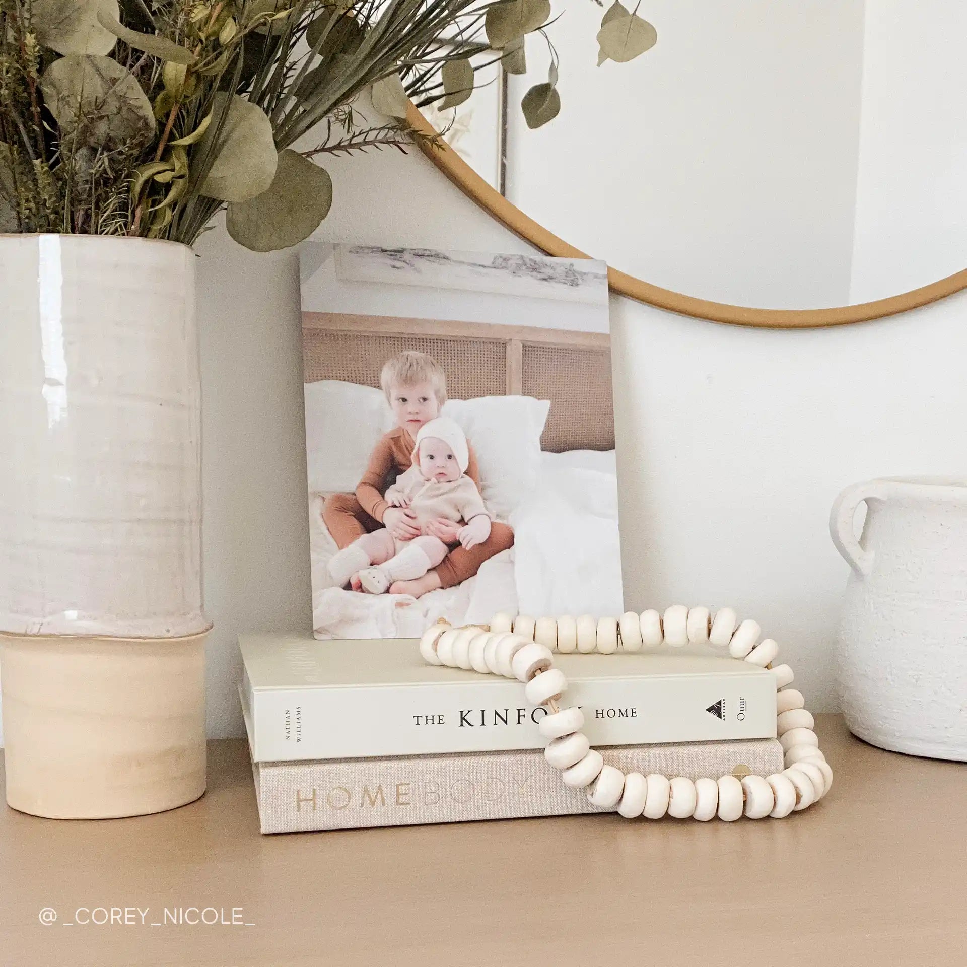 Metal wall print featuring two young children sitting together on a bed in a softly lit bedroom, displayed on stacked books beneath a round mirror.