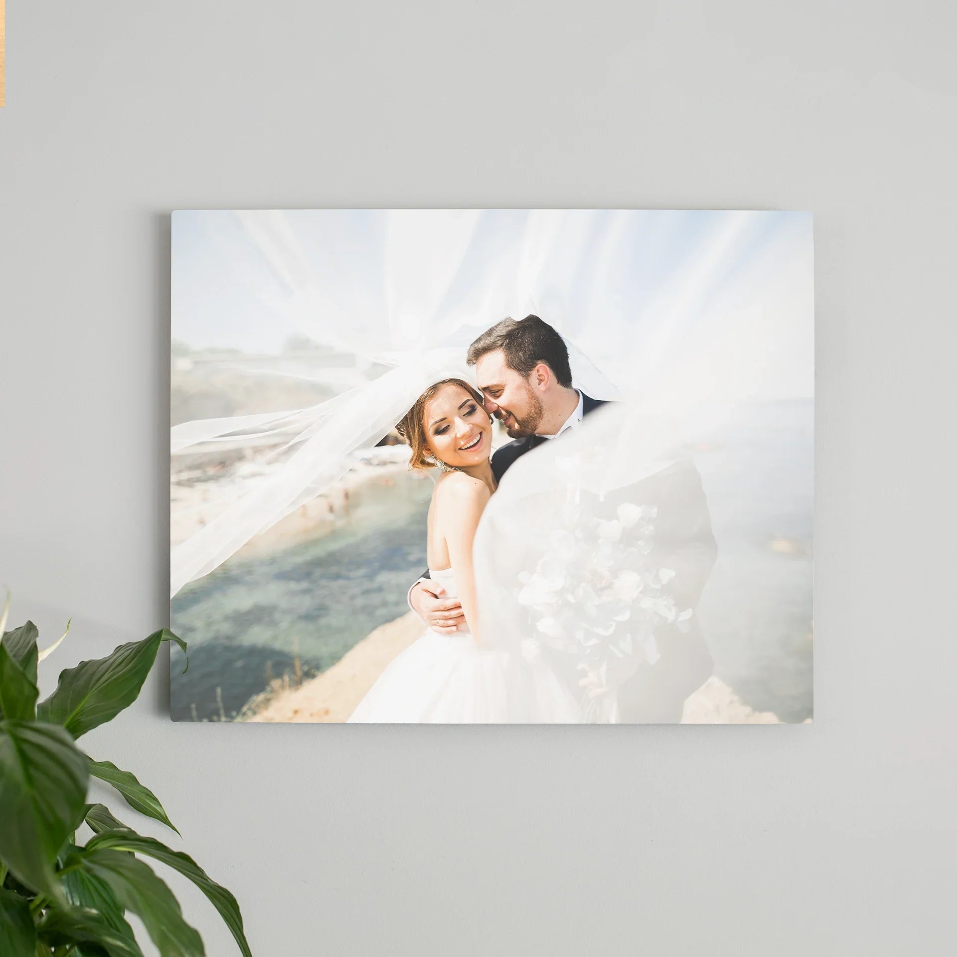 Metal wall print featuring a bride and groom embracing near a coastal cliff with flowing veil, mounted on a light gray interior wall.