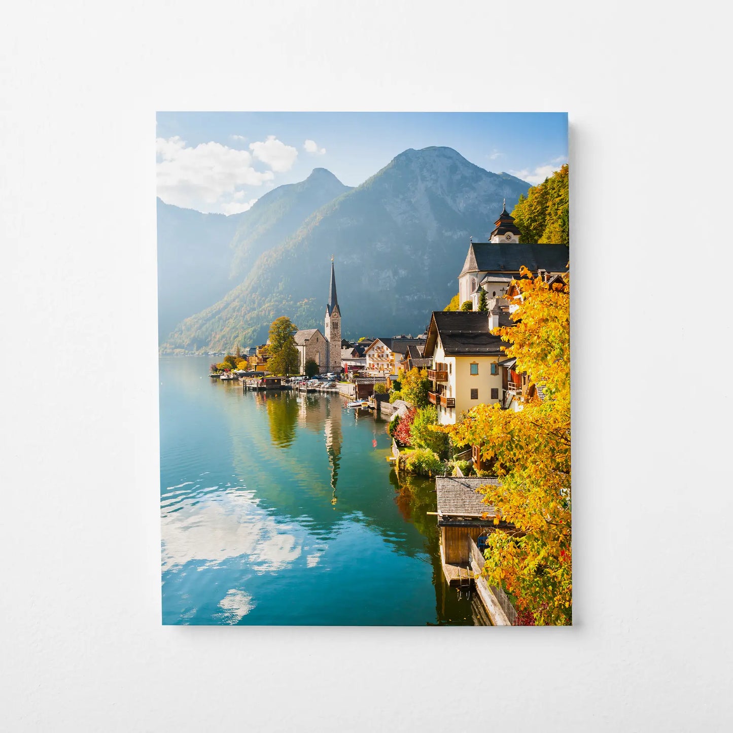 Mounted print featuring scenic lakeside village with mountains and autumn foliage, reflective water, displayed on wall.