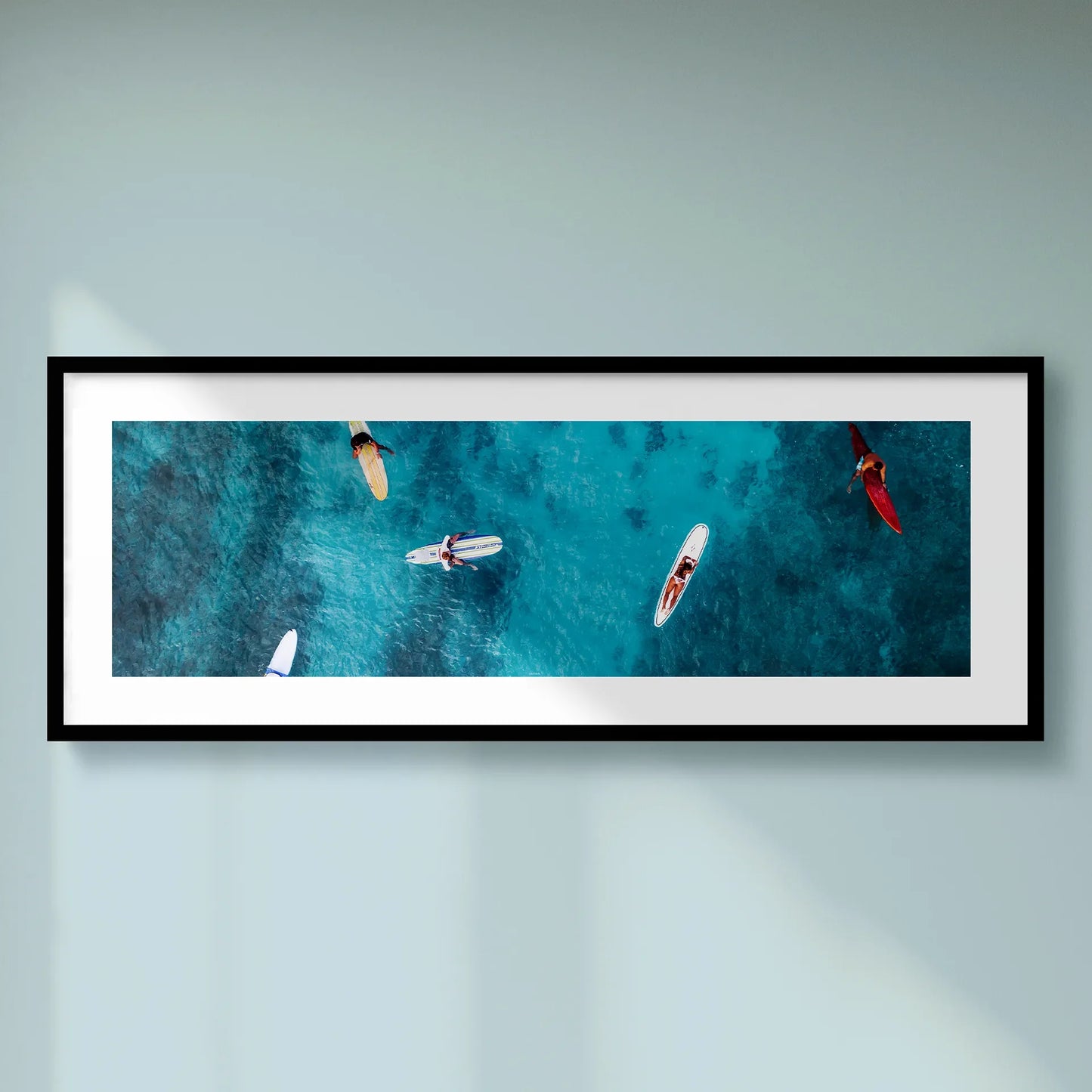 Horizontal panoramic print featuring an aerial view of surfers on turquoise ocean water, framed in black and mounted on a soft blue wall.