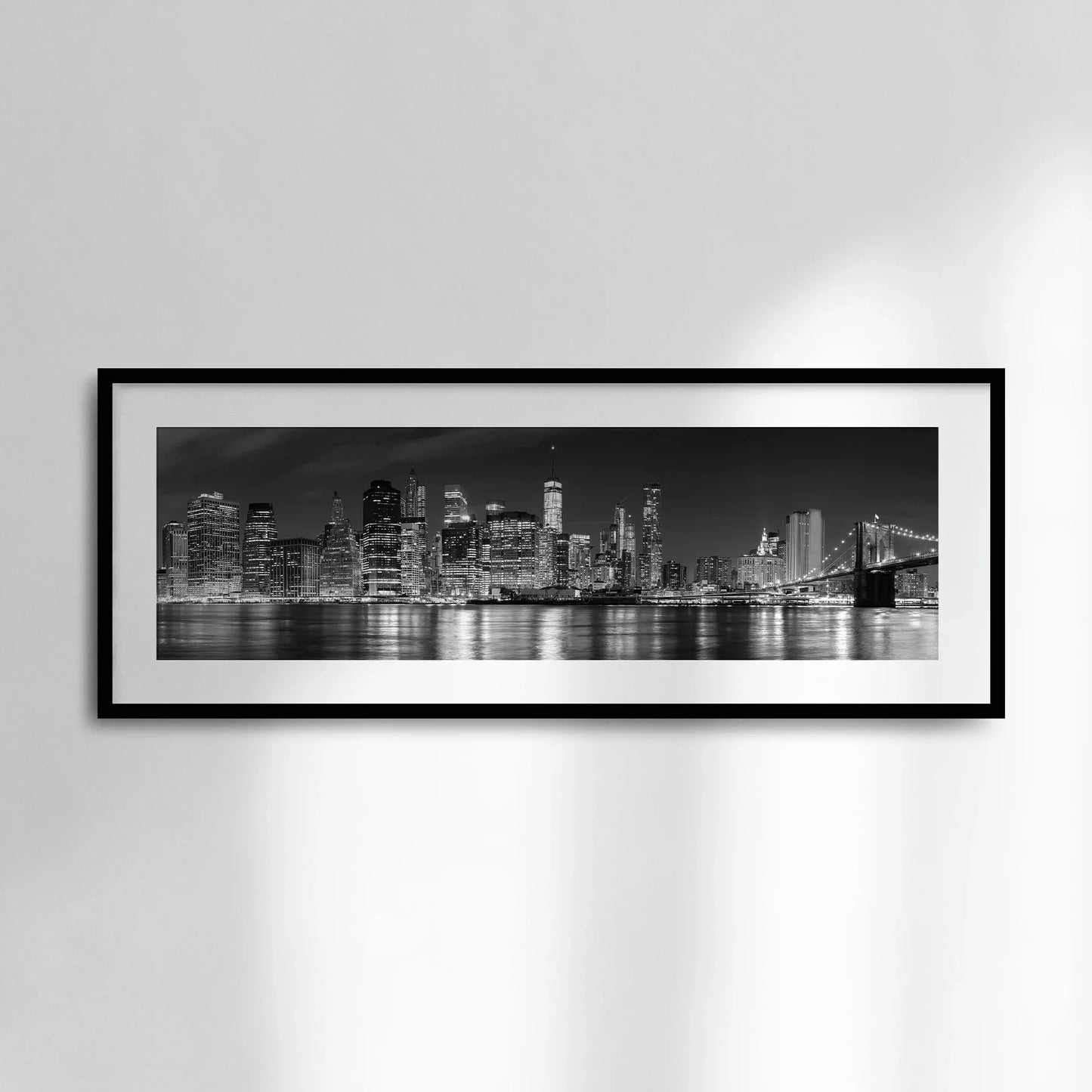 Horizontal panoramic print of a black and white city skyline with illuminated skyscrapers and bridge reflections, framed in black and displayed on a white wall.