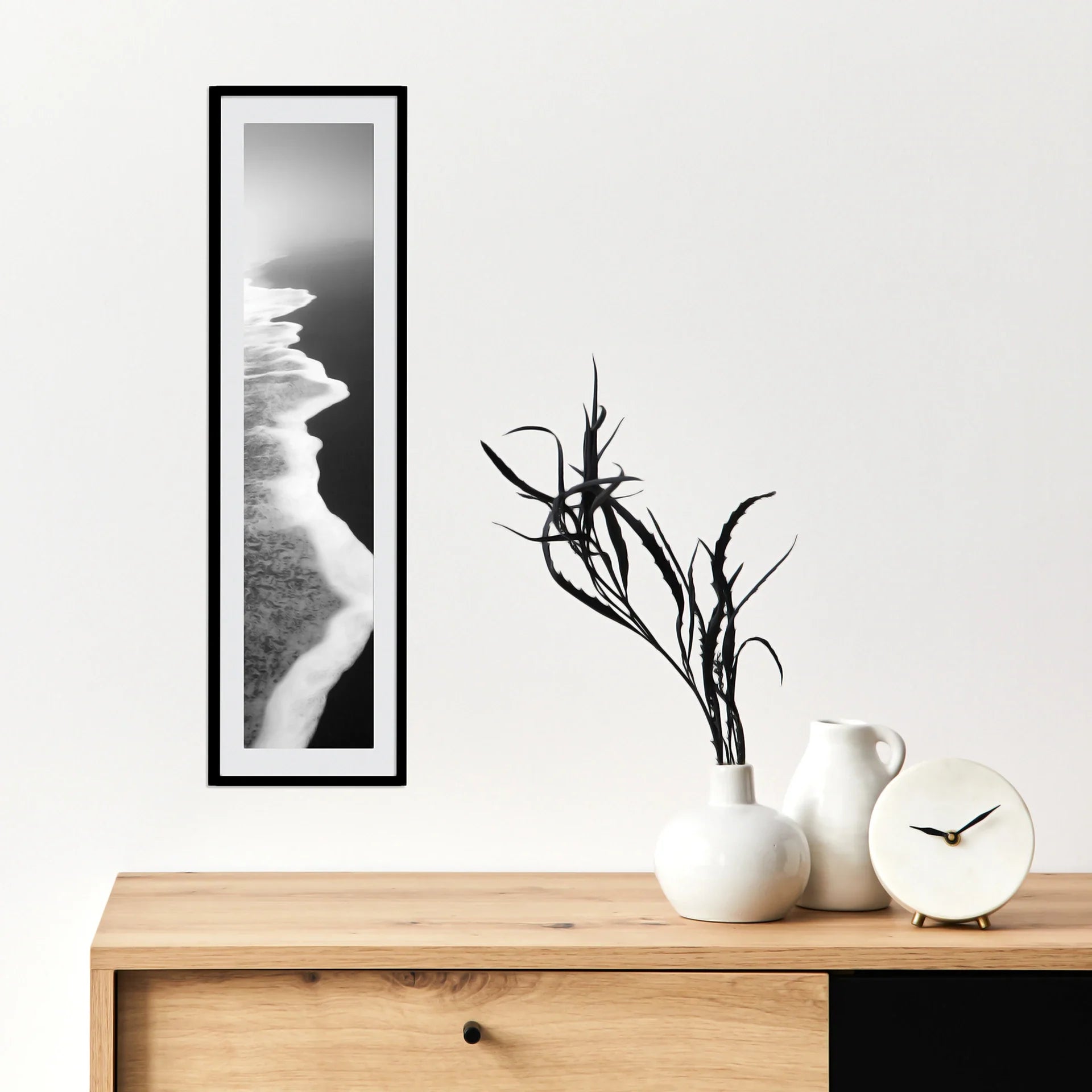Vertical panoramic print of a black and white aerial shoreline with waves meeting sand, displayed in a slim black frame above a wooden cabinet.