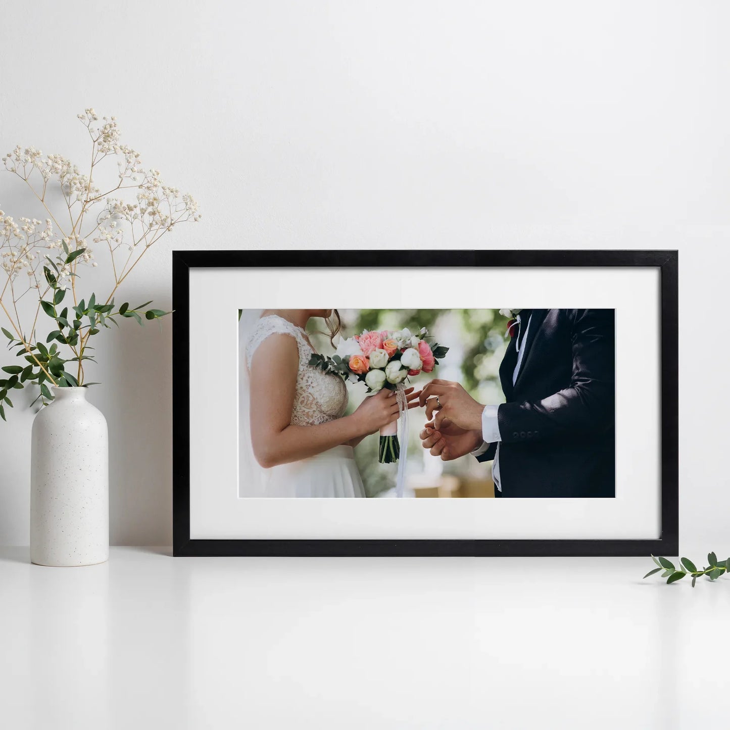 Framed panoramic photo print with white mat showing a close-up wedding moment with bouquet and hands, displayed on a tabletop with floral decor.