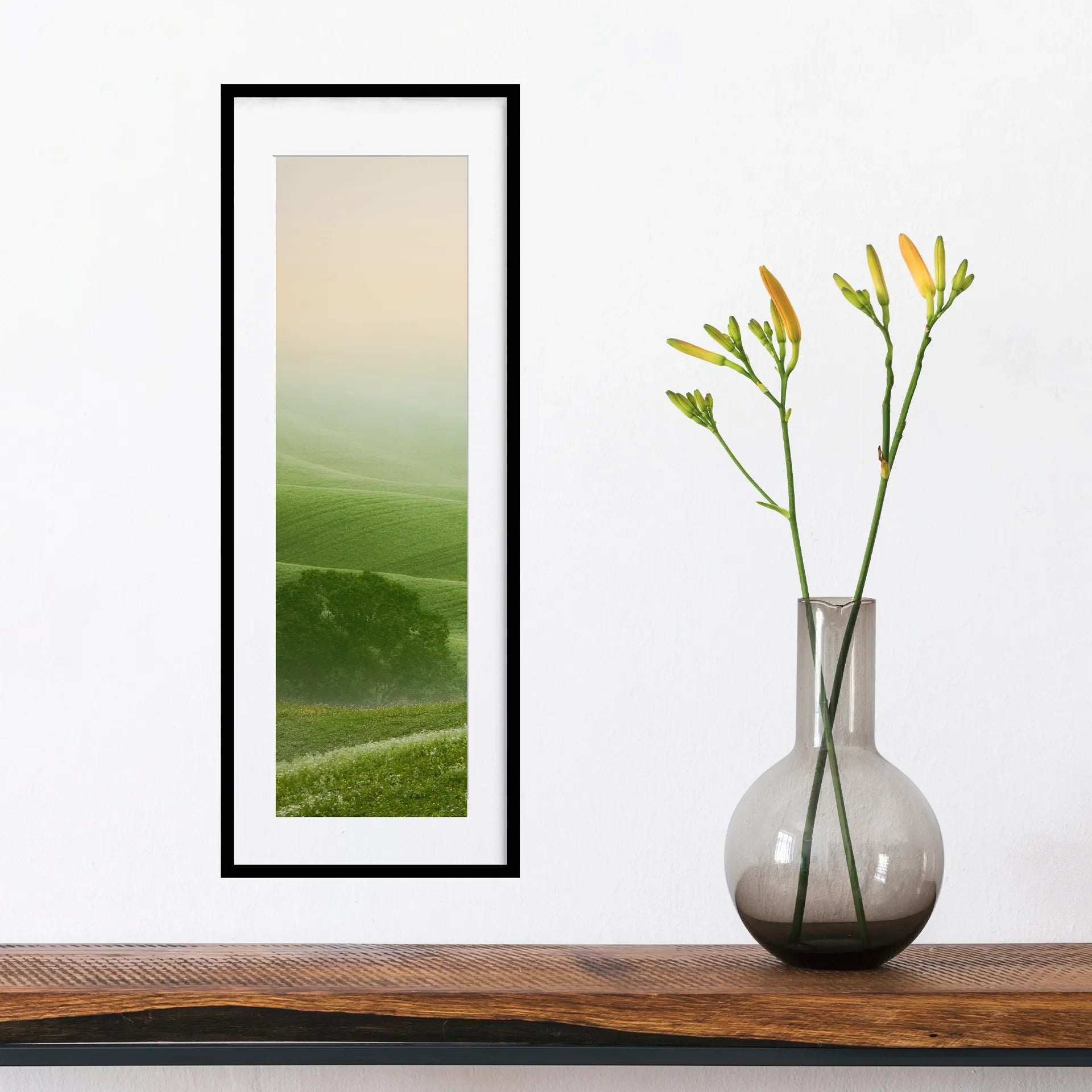 Vertical panoramic print of soft green rolling hills fading into mist, framed in black and styled above a wooden surface beside a glass vase with flowers.