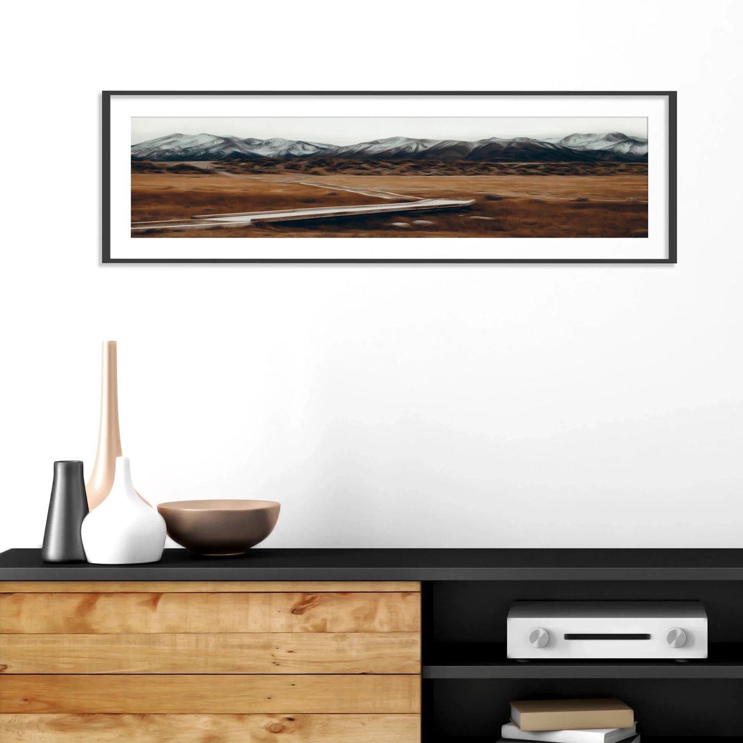 Framed panoramic print of a winding road through an open field with snow-capped mountains in the distance, mounted above a modern wood and black console.